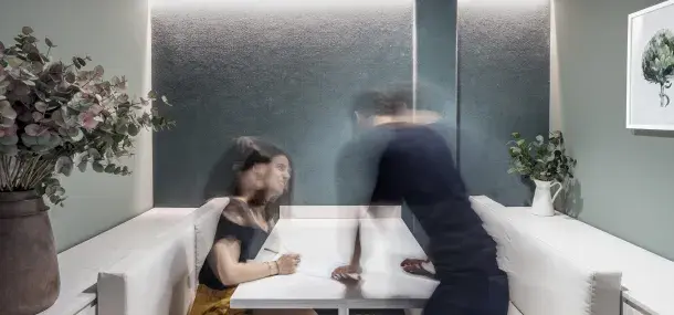 Una mujer sentada en un banco mezcla observa a un hombre desenfocado colocarse en la mesa en un moderno comedor con paredes verdes y plantas decorativas.