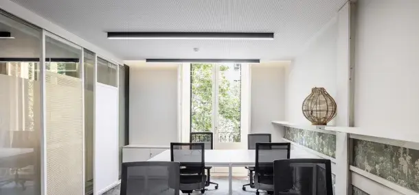 Mesa con sillas rodeándola, situada en una oficina con ventanas grandes y una lámpara decorativa sobre una repisa.paredes blancas.
