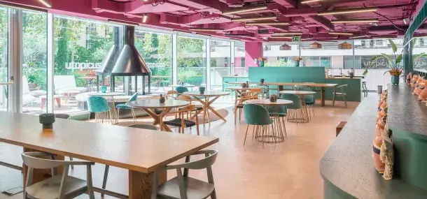 Un restaurante con mesas de madera y sillas verdes, ubicado junto a una ventana grande. En el techo, pintura rosa. Al fondo, un cuarto de estar al aire libre.