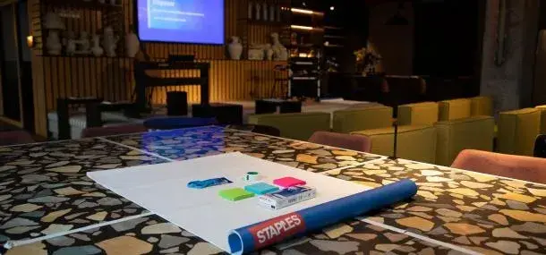 Papel y útiles de oficina sobre mesa con diseño de mosaico; pantalla de proyección y sillas en sala.