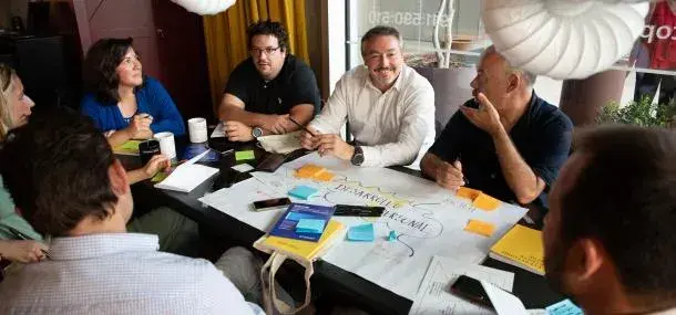 Personas sentadas conversando alrededor de una mesa, con papeles y notas adhesivas. La mesa está en un entorno interior, posiblemente una reunión de trabajo o taller colaborativo.