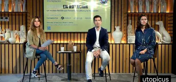 Tres personas sentadas en taburetes, participan en un panel. Detrás, un letrero dice: "EN ROTACIÓN Un modelo flexible que fomenta la cultura y la innovación". Ambiente decorado con jarrones blancos.