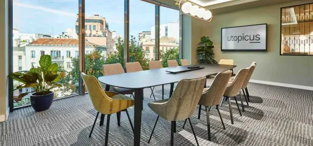 Una mesa de conferencias grande rodeada de sillas modernas se sitúa en una sala con ventanas grandes. En la pared, un monitor muestra el texto "utopicus".