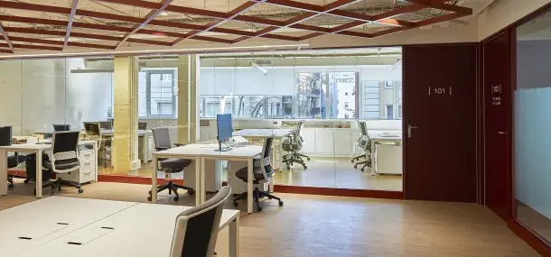 Oficina vacía con escritorios y sillas, rodeada de paneles de vidrio. En la puerta, numeración "101". Ambiente moderno con techos abiertos.
