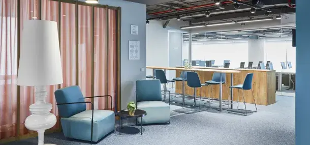 Una lámpara alta junto a sillones azules rodean una mesa pequeña. Al fondo, una barra de madera con sillas azules en un entorno de oficina moderna.