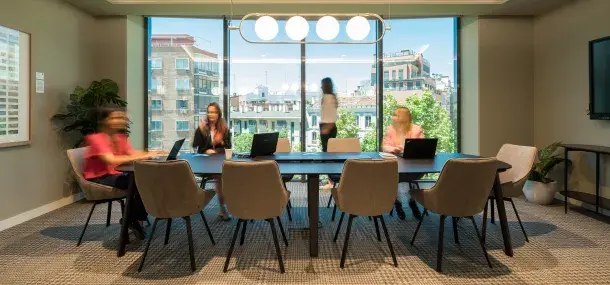 Varias personas están reunidas alrededor de una mesa de conferencias, utilizando portátiles; al fondo, una gran ventana muestra edificios y un cielo despejado.