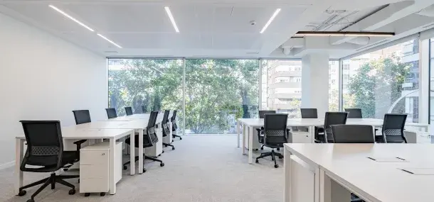 Oficinas vacías con escritorios blancos y sillas negras, frente a ventanales que muestran edificios y árboles afuera.