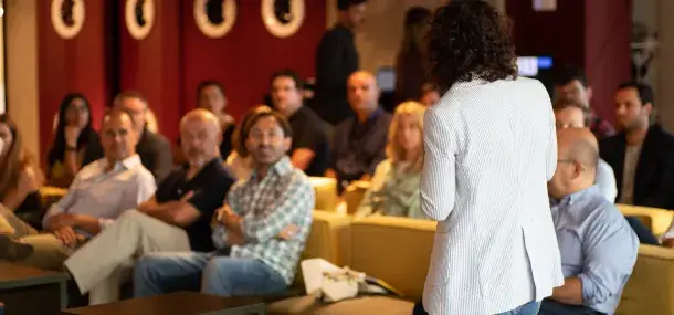 Persona de espaldas hablando frente a una audiencia atenta, sentada en sofás dentro de un salón iluminado.