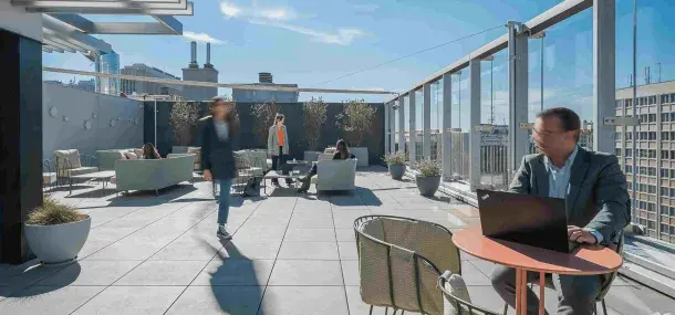 Persona sentada trabajando en una computadora portátil en una terraza soleada; otras personas socializan en sofás rodeados de plantas y edificios urbanos.