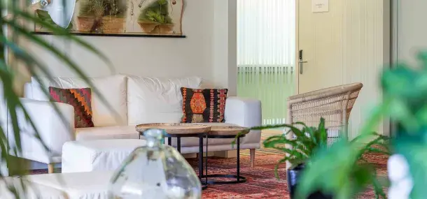 Sofá blanco con cojines coloridos, mesas de centro de madera, plantas verdes, en una sala luminosa con alfombra.