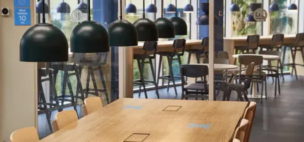 Mesa de madera clara con sillas alrededor, situada en comedor moderno; lámparas negras colgantes iluminan el espacio. En la pared, cartel azul: "aforo máximo 10 personas".