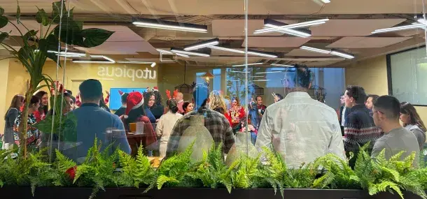 Personas están reunidas celebrando en una oficina decorada festivamente, observadas a través de una pared de vidrio. Sobre el vidrio está el texto "utópicus".