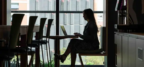 Persona trabajando en una computadora portátil, sentada en una mesa de cafetería con ventanales grandes al fondo.