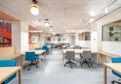 Espacio de oficina moderno con escritorios de madera y sillas giratorias. Está iluminado por lámparas colgantes, rodeado de paredes blancas y señalización de emergencia roja en un rincón.
