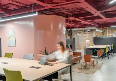 Persona desdibujada gesticula en una mesa con portátil y teléfono, en oficina moderna con paredes rosadas.