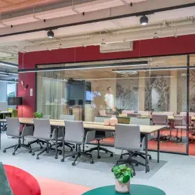 Un espacio moderno con sillones rojos y grises, personas conversan y trabajan en un entorno luminoso. Decoración contemporánea y plantas. Letreros visibles: "SALIDA" y un panel con pantalla iluminada.